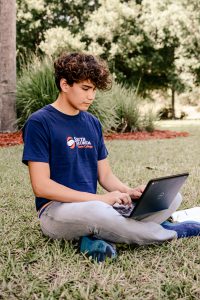 Male student with curly hair sitting on the lawn and typing on a laptop.