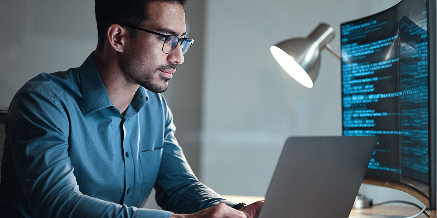 A man with glasses works on a laptop with a second monitor showing code
