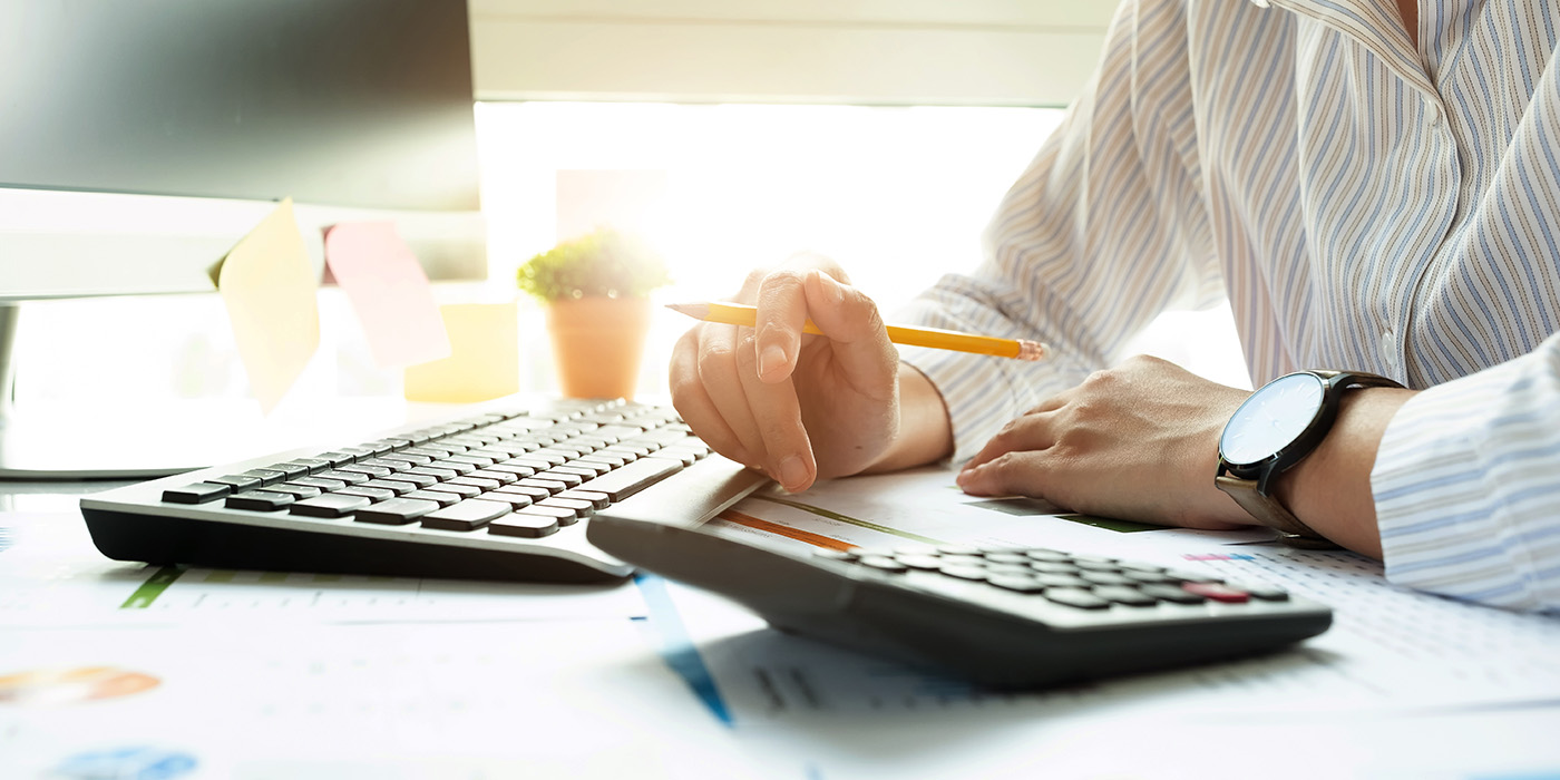 Person working at a desk with a computer, calculator, and financial documents, analyzing data with a pencil in hand.