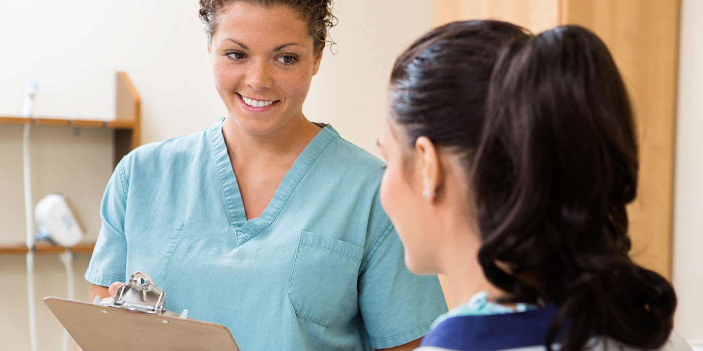 A nurse in scrubs smiling and holding a clipboard while speaking with a patient in a medical office