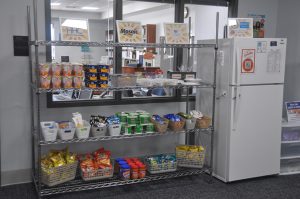 A shelving unit with food on each shelf and the logos of supporters on the top shelf. A refrigerator is set up next to the shelving unit. 
